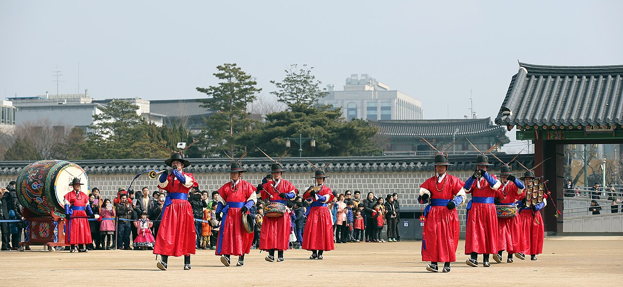 Performers in red court costumes play drums and horns in a Seoul palace courtyard as spectators watch